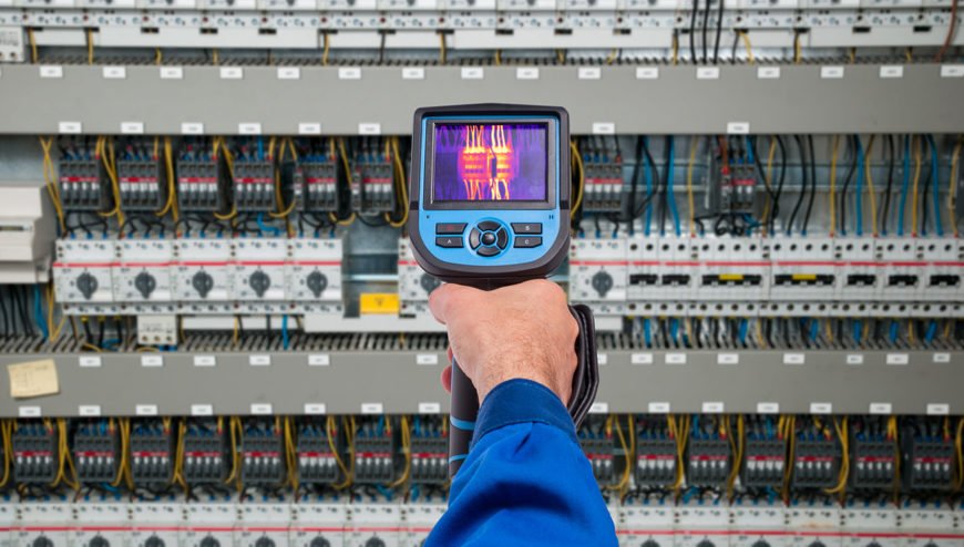 Técnico utilizando uma câmera termográfica para operar um painel elétrico. A tela da câmera exibe um termograma com variações de cor que mostram diferentes temperaturas em componentes elétricos, destacando áreas de calor que podem sinalizar possíveis falhas ou anomalias no sistema. Uma imagem exemplifica a aplicação prática da termografia elétrica na manutenção preditiva.